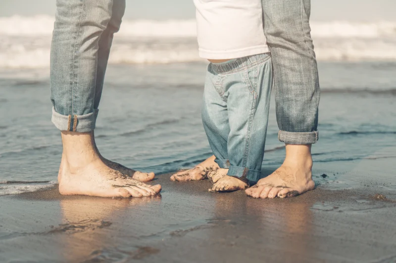 Fotografía en la que se ven los pies de tres personas en la playa: una mujer, un hombre y un bebé.